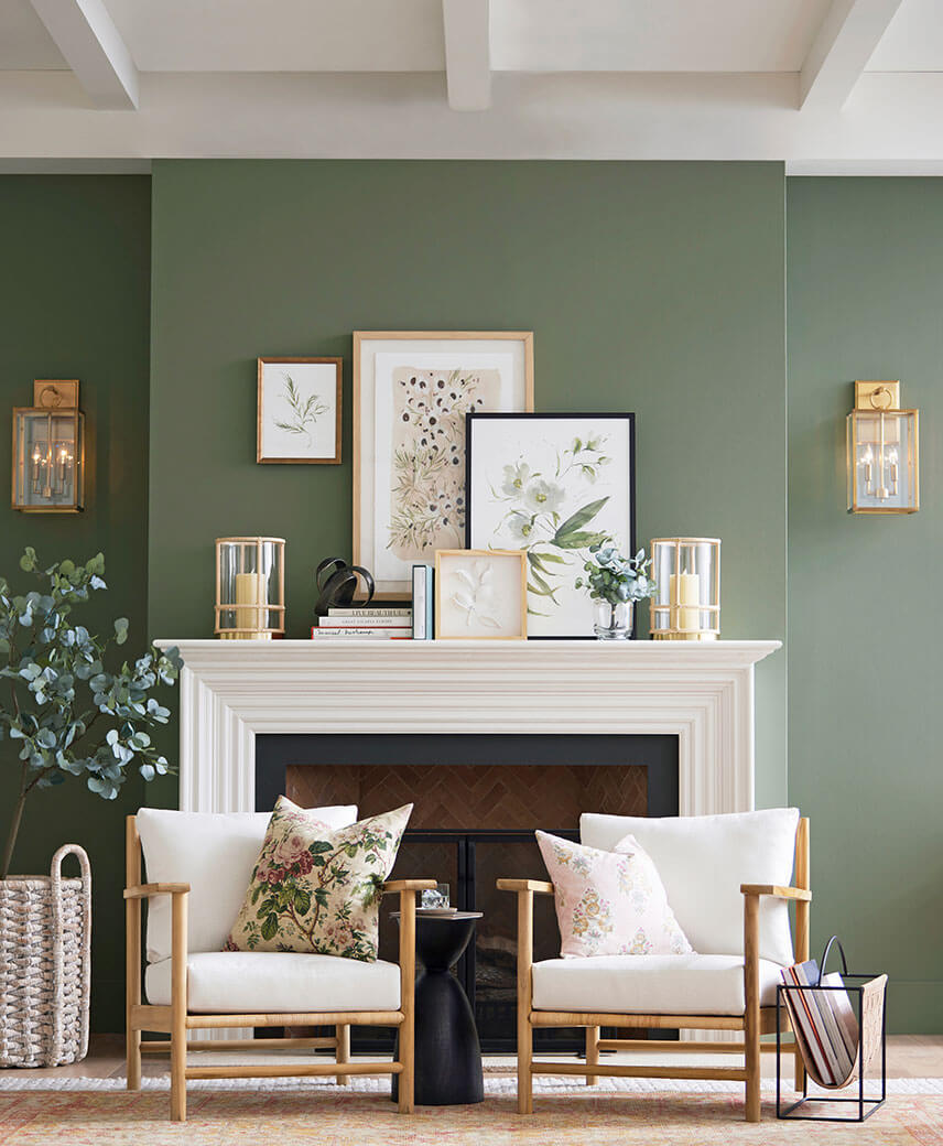 Pottery Barn living room with walls painted in Dried Thyme SW 6186, large fireplace with the mantle decorated with art, candles, books, and two matching chairs in front.